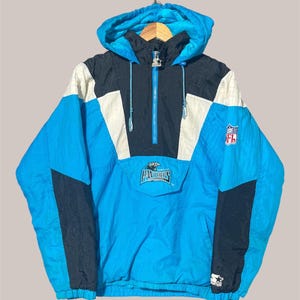 May include: A Carolina Panthers Starter pullover hooded jacket in bright blue, black, and white. The jacket features a half-zip front, a front pouch pocket with the Panthers logo, an NFL shield patch on the sleeve, and a Starter logo on the cuff.