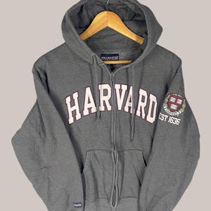 以下が含まれることがあります： 胸に白いブロック体で「HARVARD」の文字が入った、グレーのジップアップパーカー。フード、フロントポケット、ハーバード大学の紋章と「EST 1636」のパッチが袖に付いています。