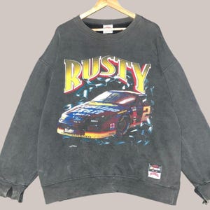 May include: A black sweatshirt with a faded graphic of a racing car and the word "RUSTY" in yellow. The car is a Ford Thunderbird with the number 2 on the side. The sweatshirt has a small tag on the bottom right corner that says "Steiner".