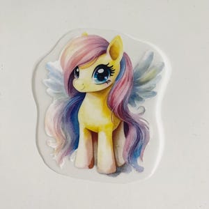 Peut inclure: Un poney jaune avec une crinière et une queue rose, violette et bleue. Le poney a des yeux bleus et des ailes blanches. Le poney est debout sur un fond blanc.