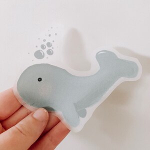 Può includere: Un adesivo di balena blu chiaro su sfondo bianco. La balena sta nuotando con bolle che escono dal suo sfiatatoio.