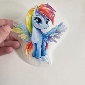 Peut inclure: Un poney de dessin animé bleu et arc-en-ciel avec des ailes. Le poney a une crinière et une queue arc-en-ciel, et ses ailes sont d'un bleu clair avec des accents arc-en-ciel.