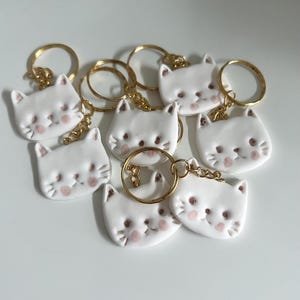 May include: A collection of white ceramic cat-shaped keychains with gold-colored key rings. Each keychain features a smiling cat face with pink blush on the cheeks. The keychains are arranged on a white surface.