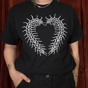 May include: Black t-shirt featuring a white heart-shaped design composed of centipede-like figures with heart details. The graphic tee has short sleeves and a crew neck. The shirt is made of a soft, comfortable material.