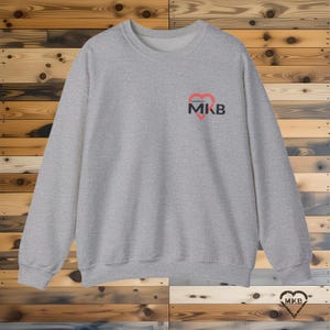 May include: A light gray sweatshirt with a crew neck. The sweatshirt features a small design on the upper left chest, consisting of a red heart shape with the letters "MKB" in black. The background is a wooden plank wall.