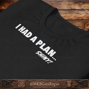 May include: Black t-shirt with white text that reads "I HAD A PLAN... SHINY!". The shirt has a crew neck and is made of a soft material. The text is angled across the front of the shirt.
