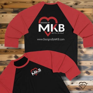 May include: A black and red raglan baseball shirt with the text "DESIGNS BY MKB Art From the Heart" and the website address www.DesignsByMKB.com. The shirt features a heart-shaped logo with the initials MKB.