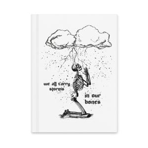 May include: White notebook with a black and white illustration of a skeleton kneeling under a storm cloud with lightning. The text reads "we all carry storms in our bones."