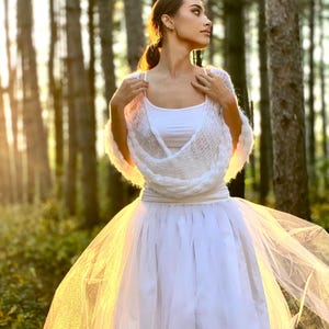 May include: A woman wearing a white crocheted shawl and a white tulle skirt stands in a forest. The sun shines through the trees, creating a warm glow.
