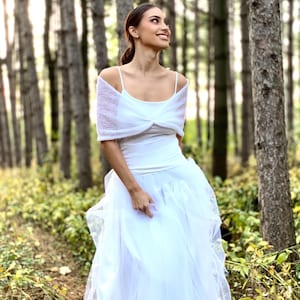 May include: A woman wearing a white dress and a white knitted shawl stands in a forest. She is smiling and looking upwards.