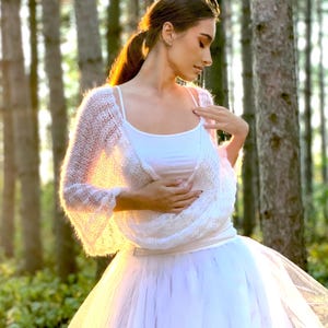 May include: A white crochet shrug with a delicate open-weave pattern. The shrug is worn over a white vest top and a white tulle skirt. The model is standing in a woodland setting.