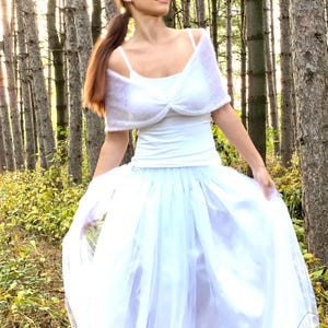 May include: A white outfit comprising a spaghetti strap top, a fluffy shrug, and a long tulle skirt. The outfit is worn outdoors in a forest setting with tall trees and sunlight.
