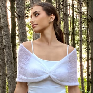 May include: A white knitted shrug with a knotted detail at the front. The shrug is worn over a white vest top. The model is standing in a woodland setting.