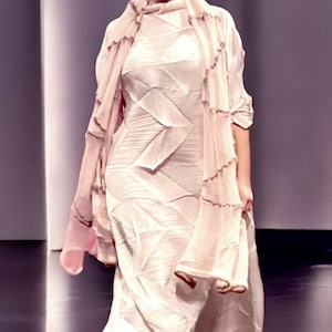 Puede incluir: Una mujer lleva un vestido largo, fluido y rosa con un patrón geométrico. El vestido tiene un diseño drapeado y en capas y una silueta suelta y fluida.