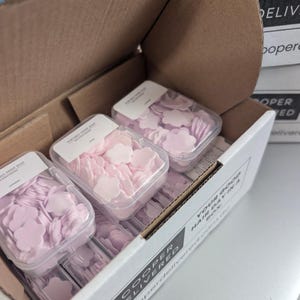 May include: A cardboard box containing three clear plastic containers of flower-shaped paper soap. The soap is in shades of pink and lavender. The box has the words "COOPER DELIVERED" printed on the side.