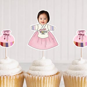 May include: Four cupcakes with white frosting, each topped with a decorative pick. Two picks feature a cartoon image of a baby in traditional Korean clothing, and two have a pink pouch design.
