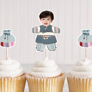 May include: Four cupcakes with white frosting, each topped with a decorative pick. Two picks feature a baby in traditional Korean clothing, and two have a colorful pouch design. The cupcakes are in yellow paper liners.
