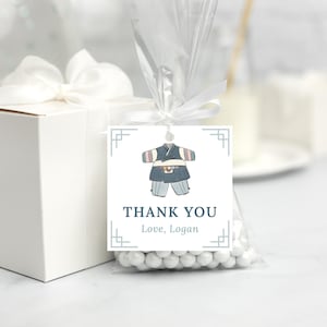 Peut inclure: Un sac cadeau blanc avec un nœud blanc et une étiquette de remerciement. L'étiquette représente une illustration bleue et rose d'une tenue de bébé coréenne traditionnelle et le texte "Merci Amour, Logan".