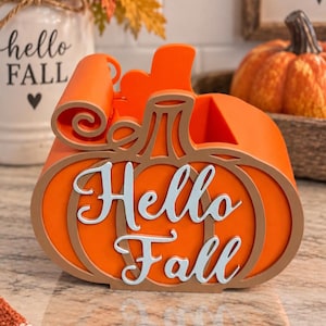 May include: An orange pumpkin-shaped desk organizer featuring a tan outline and the white script text Hello Fall on the front. The container has a hollow interior space and a curved top edge, constructed with layered panels in a bright orange finish.