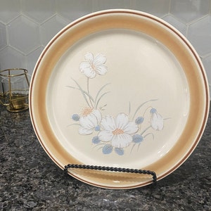 Puede incluir: Un plato de cena de cerámica marrón y blanco con un diseño floral. El plato presenta un borde marrón y un centro blanco con un patrón floral de flores blancas y hojas verdes.