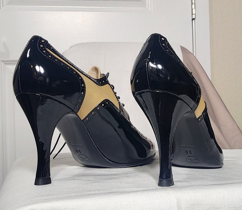 K&ouml;nnte beinhalten: Ein Paar schwarze Lacklederschuhe mit Absatz, braunem Lederbesatz und Schn&uuml;rung. Die Schuhe haben einen hohen Absatz und eine spitze Zehenpartie.