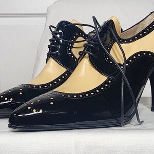K&ouml;nnte beinhalten: Schwarze und beigefarbene Lederschuhe mit Absatz, Schn&uuml;rung vorne und Brogue-Muster. Die Schuhe haben eine spitze Zehenpartie und einen hohen Absatz.