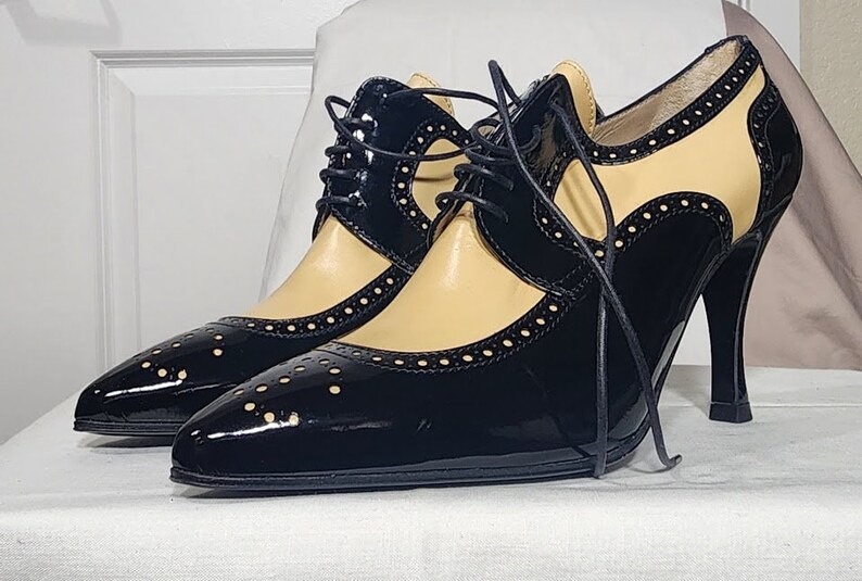 K&ouml;nnte beinhalten: Schwarze und beigefarbene Lederschuhe mit Absatz, Schn&uuml;rung vorne und Brogue-Muster. Die Schuhe haben eine spitze Zehenpartie und einen hohen Absatz.