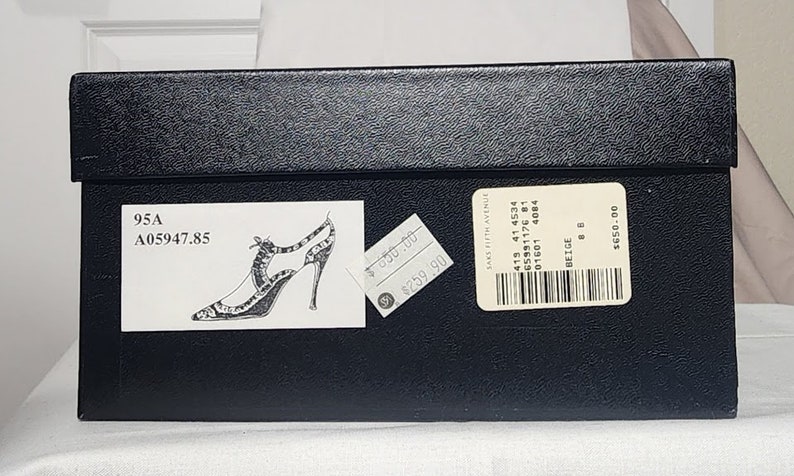 K&ouml;nnte beinhalten: Ein schwarzer Schuhkarton mit einem wei&szlig;en Etikett, auf dem "95A A05947.85" steht und eine Zeichnung eines schwarz-wei&szlig;en High-Heel-Schuhs. Es gibt ein Preisschild mit der Aufschrift "259,90 &euro;" und ein wei&szlig;es Etikett mit Barcode und Text, auf dem "SAKS FIFTH AVENUE 415 41 4534 65991176 81 01601 4084 BEIGE 8 650,00 &euro;" steht.