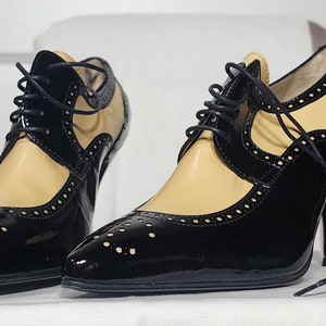 K&ouml;nnte beinhalten: Ein Paar schwarze und braune Lederschuhe mit Schn&uuml;rung und Brogue-Muster. Die Schuhe haben eine spitze Zehenpartie und einen hohen Absatz.
