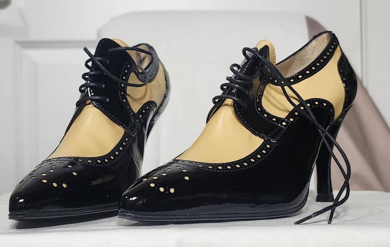 K&ouml;nnte beinhalten: Ein Paar schwarze und braune Lederschuhe mit Schn&uuml;rung und Brogue-Muster. Die Schuhe haben eine spitze Zehenpartie und einen hohen Absatz.