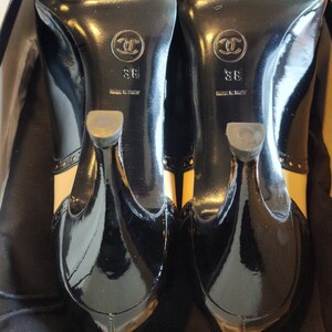 K&ouml;nnte beinhalten: Ein Paar schwarze Lackleder-Schuh mit Absatz, beigefarbener Verzierung und einem kleinen goldenen Chanel-Logo auf der R&uuml;ckseite jedes Schuhs. Die Schuhe sind Gr&ouml;&szlig;e 38 und in Italien hergestellt.