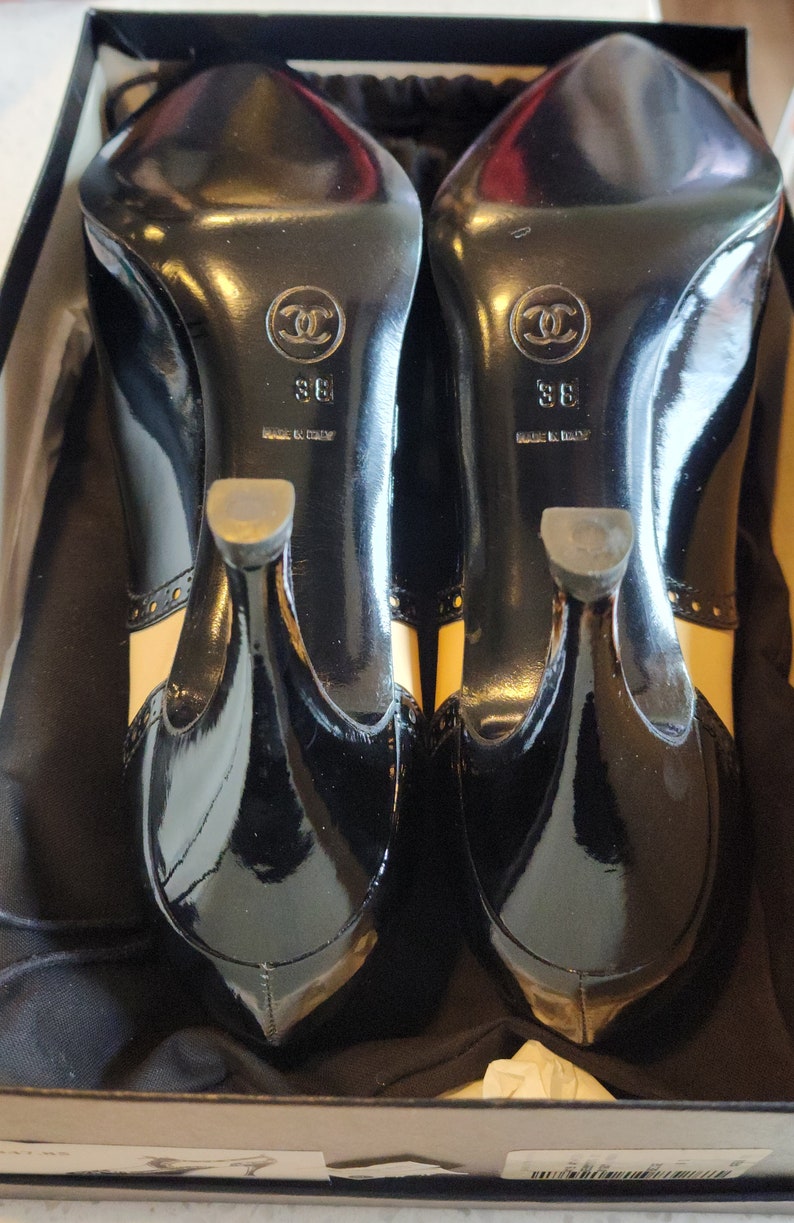 K&ouml;nnte beinhalten: Ein Paar schwarze Lackleder-Schuh mit Absatz, beigefarbener Verzierung und einem kleinen goldenen Chanel-Logo auf der R&uuml;ckseite jedes Schuhs. Die Schuhe sind Gr&ouml;&szlig;e 38 und in Italien hergestellt.