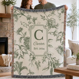 May include: A woven throw blanket featuring a cream background with a green botanical floral pattern. The centre displays a rectangular frame containing the letter C, the name Carter, and the text EST. 2024. The blanket has fringed edges.