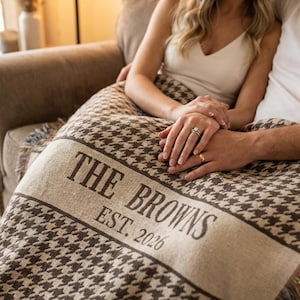 Puede incluir: Una manta marrón y crema con estampado de pata de gallo con el texto "THE BROWNS EST. 2026" en una fuente serif. La manta tiene un borde con flecos y está sobre un sofá.