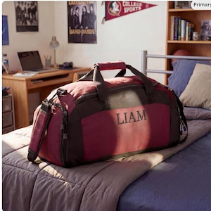 May include: A maroon and black duffel bag with the name "LIAM" embroidered on the front. The bag has a top handle and a shoulder strap. It is sitting on a bed with a gray comforter and blue sheets.
