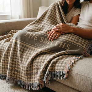May include: A woven throw blanket featuring a brown and beige houndstooth pattern with a central horizontal band. The text THE SMITHS EST. 2026 is printed in a light beige font across the band. The blanket has fringed edges on the bottom.