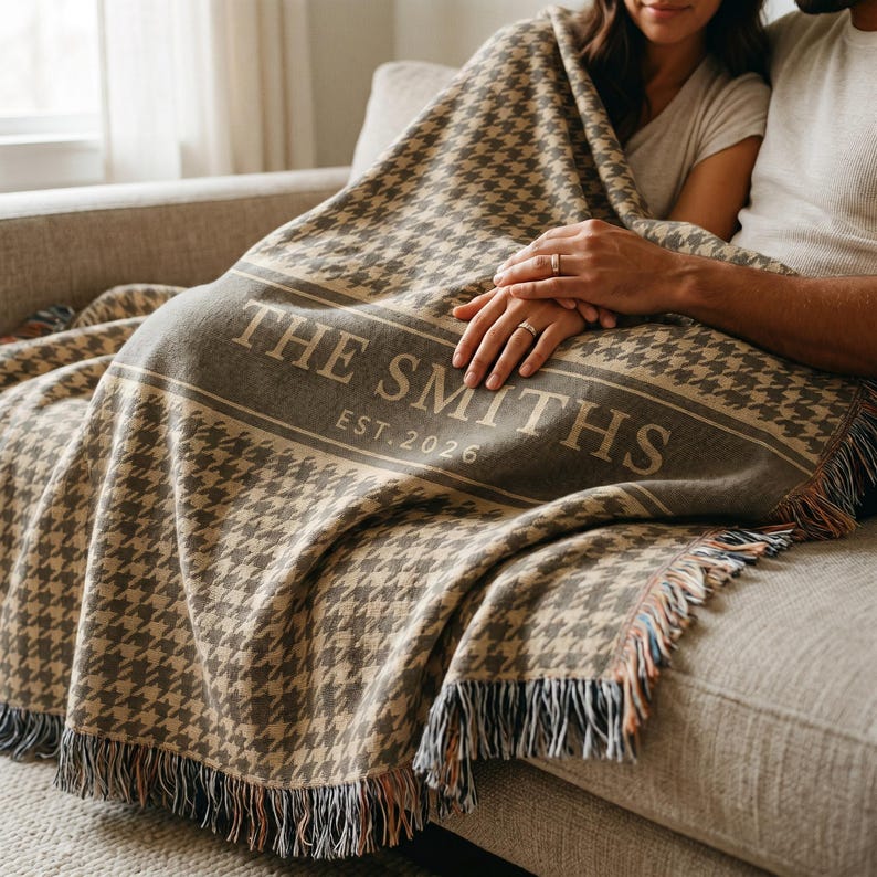 May include: A woven throw blanket featuring a brown and beige houndstooth pattern with a central horizontal band. The text THE SMITHS EST. 2026 is printed in a light beige font across the band. The blanket has fringed edges on the bottom.