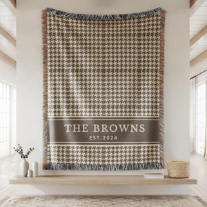 May include: A rectangular woven throw blanket featuring a brown and cream houndstooth pattern. A solid brown horizontal band across the center displays the white text THE BROWNS and EST. 2024. The blanket has fringed edges with multicolored threads.