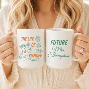 May include: Two white ceramic mugs. The left mug features orange and green text reading THE LIFE OF A FIANCEE with illustrations of a disco ball, cake, shoes, ring, champagne, and wedding banner. The right mug has green text reading FUTURE Mrs. Thompson.