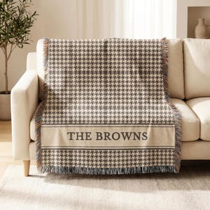 May include: A woven throw blanket featuring a brown and cream houndstooth pattern. The blanket has a horizontal band near the bottom with the text THE BROWNS in dark brown capital letters, framed by thin lines, and finished with fringed edges.