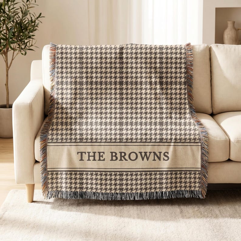May include: A woven throw blanket featuring a brown and cream houndstooth pattern. The blanket has a horizontal band near the bottom with the text THE BROWNS in dark brown capital letters, framed by thin lines, and finished with fringed edges.