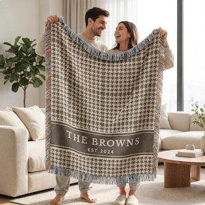 May include: A woven throw blanket featuring a brown and cream houndstooth pattern with a solid horizontal band containing the white text THE BROWNS and EST. 2024. The blanket has a fringed edge with multicolored threads along the vertical sides.