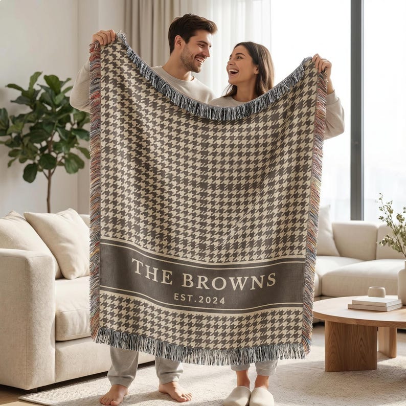 May include: A woven throw blanket featuring a brown and cream houndstooth pattern with a solid horizontal band containing the white text THE BROWNS and EST. 2024. The blanket has a fringed edge with multicolored threads along the vertical sides.