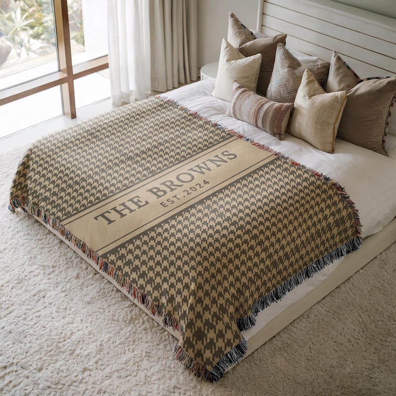 May include: A woven throw blanket featuring a brown and beige houndstooth pattern with a central horizontal band. The band displays the text THE BROWNS and EST. 2024 in dark lettering. The blanket has fringed edges along the top and bottom sides.