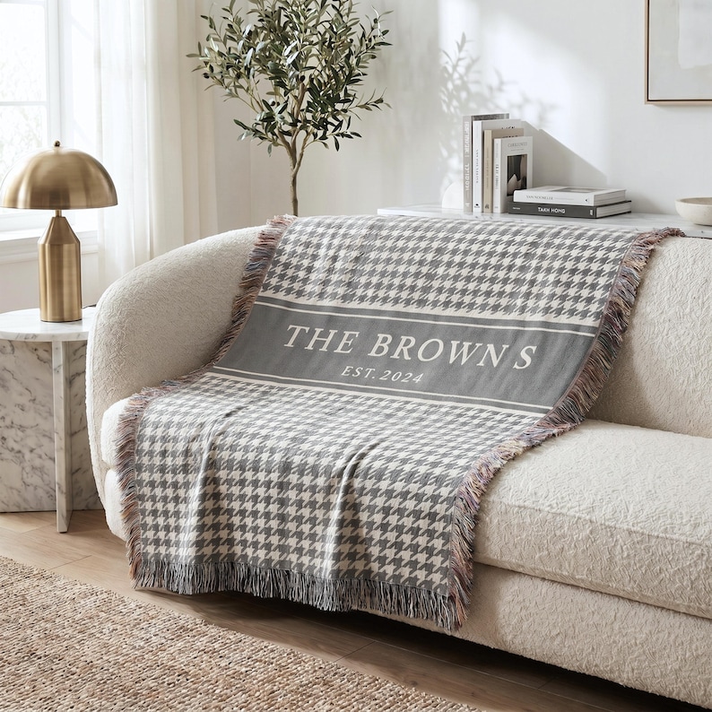 May include: A woven throw blanket featuring a grey and white houndstooth pattern with a solid grey horizontal band across the center. The text THE BROWNS and EST. 2024 are printed in white on the band. The blanket has fringed edges on the sides.
