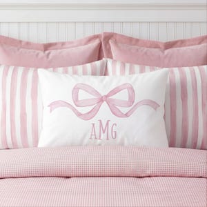 May include: A white pillow with a watercolor bow and the letters "AMG" in pink. The pillow is on a bed with pink and white striped pillows and a pink patterned bedspread. The bedding has a soft, feminine aesthetic.