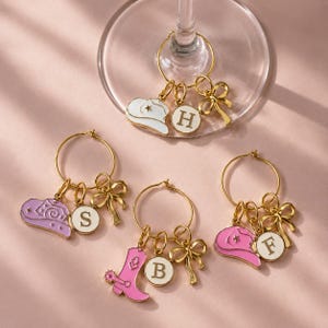 May include: Gold-toned wine charms with cowboy hat, boot, and bow designs. Charms include white, pink, and purple elements, with initial letters on round tags. The charms are hanging on a wine glass.