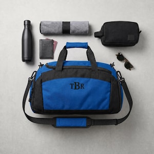 Könnte beinhalten: Draufsicht auf Reiseaccessoires, darunter eine schwarz-blaue Reisetasche mit gesticktem "TBr", eine schwarze Wasserflasche, ein graues Handtuch, eine schwarze Kulturtasche, Sonnenbrille und ein Reisepasshalter.