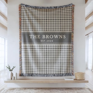 May include: A rectangular woven throw blanket featuring a grey and white houndstooth pattern. A solid grey horizontal band across the center displays the white text THE BROWNS and EST. 2024. The blanket has fringed edges with multicolored threads.