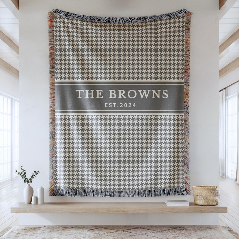 May include: A rectangular woven throw blanket featuring a grey and white houndstooth pattern. A solid grey horizontal band across the center displays the white text THE BROWNS and EST. 2024. The blanket has fringed edges with multicolored threads.